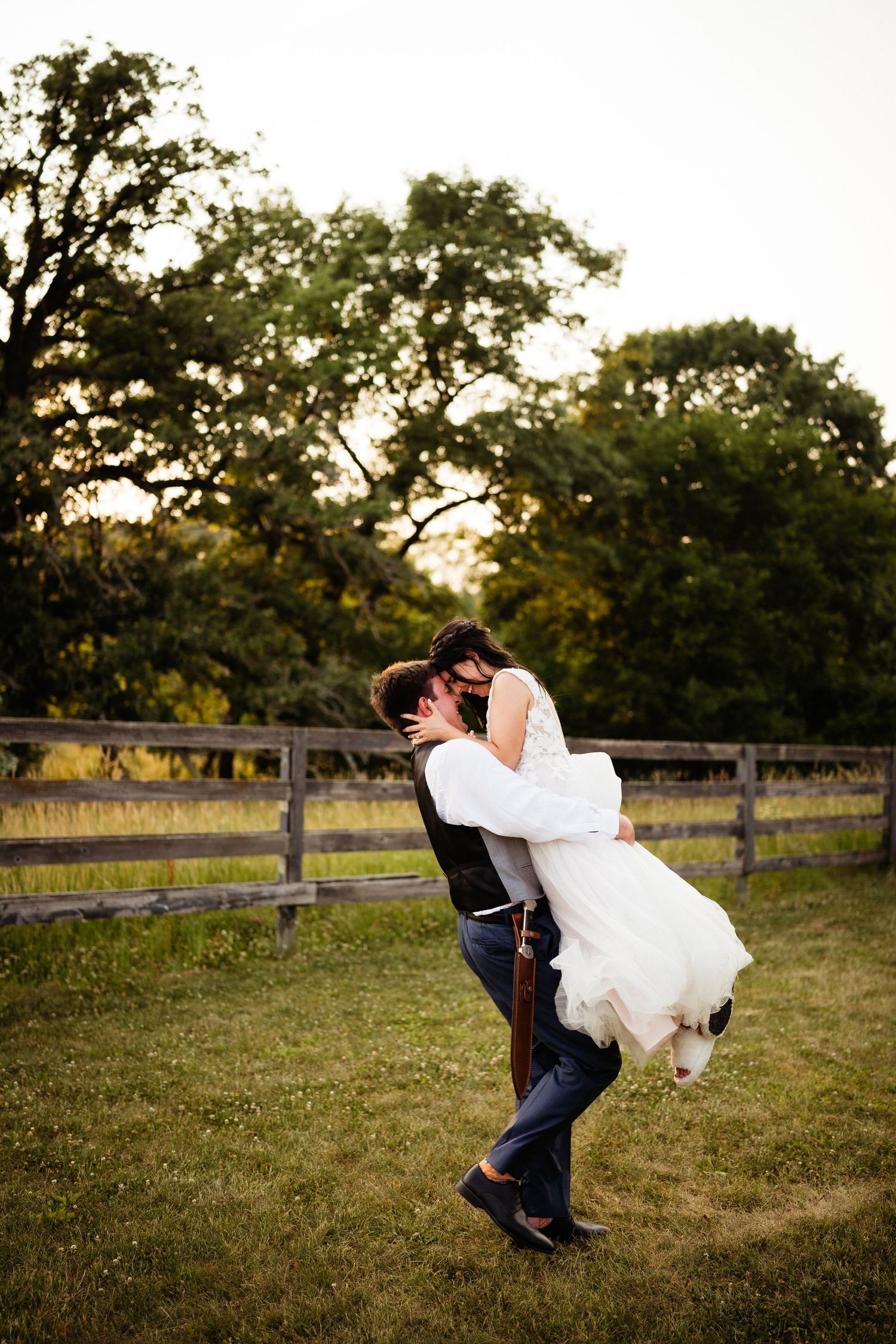 A romantic wedding couple shares a playful moment in a rustic outdoor setting with wooden fences and trees at sunset.