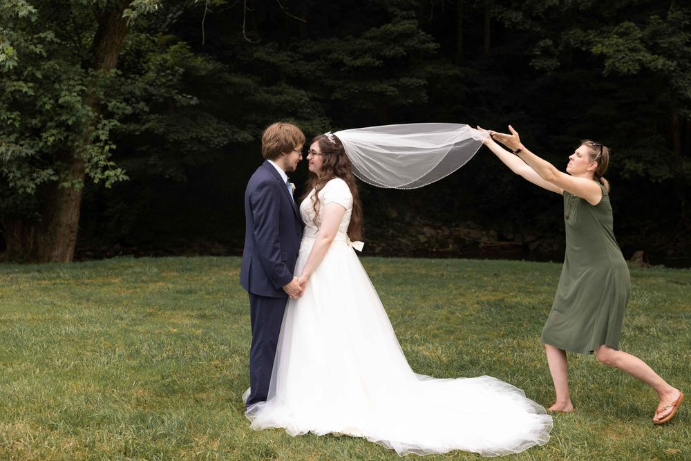 Wedding couple embraces on grass while veil billows dramatically in summer breeze.