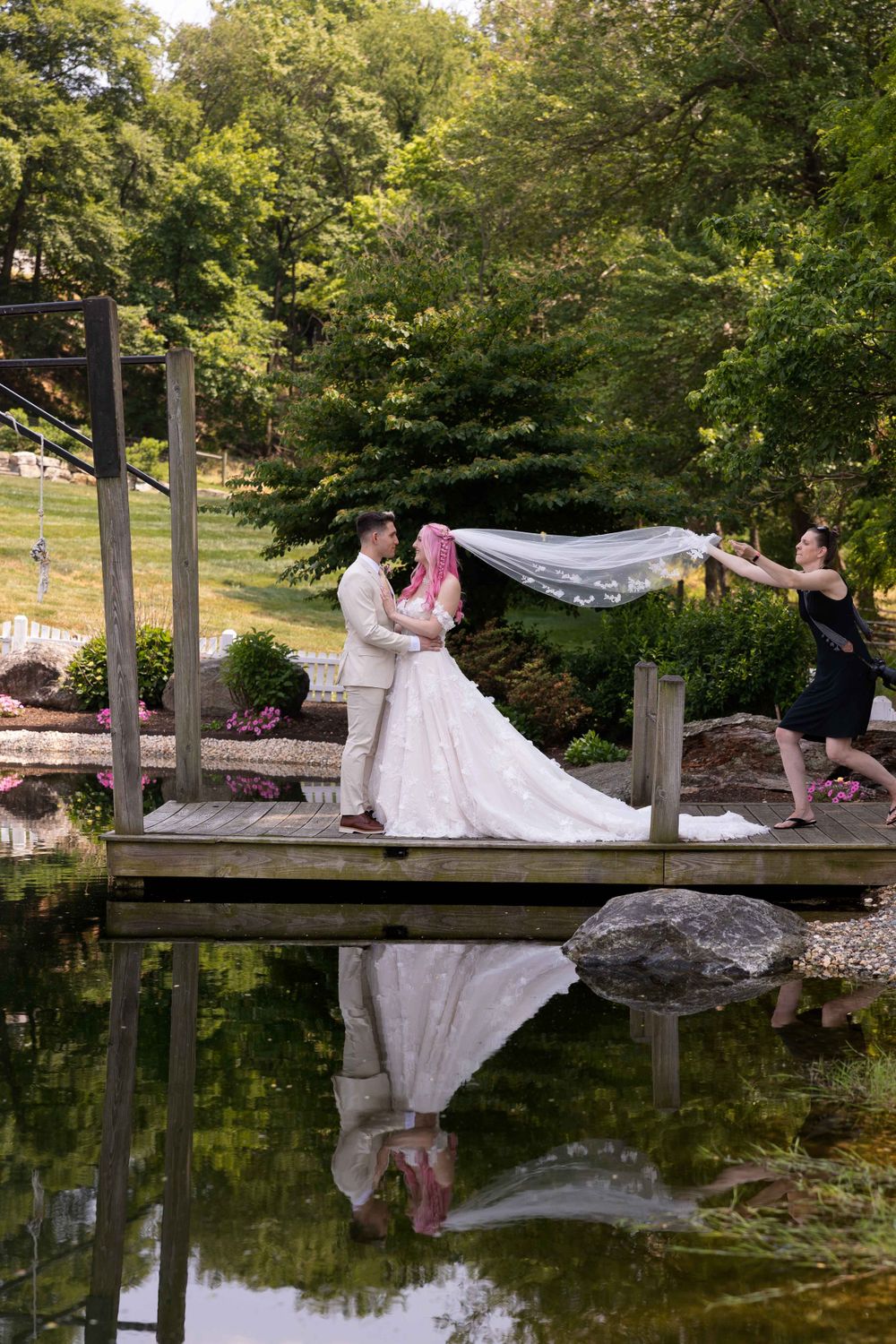 Elegant wedding portrait on wooden bridge over pond with veil flowing in reflection.