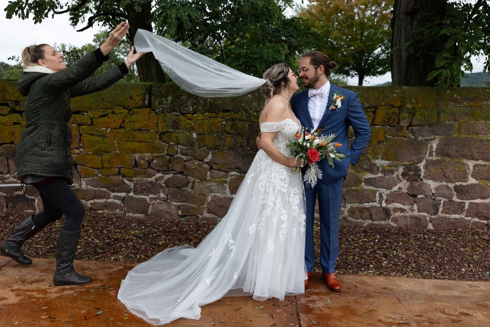Wedding photo against stone wall with flowing veil caught in autumn wind.