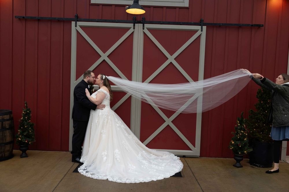 Wedding couple poses against red barn door while veil flows dramatically in breeze.