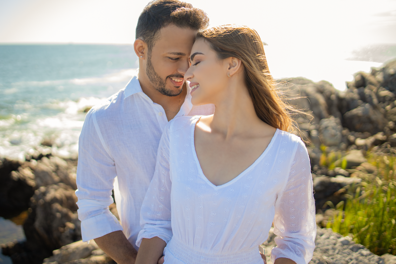 Couple in white clothing embraces on rocky coastline with ocean view.