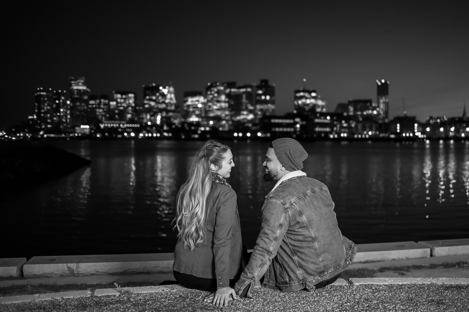 A couple sits together by the water at night with city lights glowing in the background across the river.