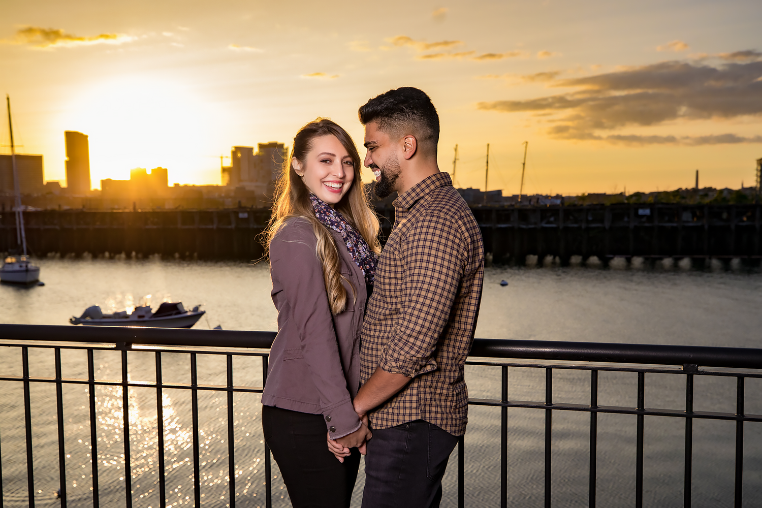 Couple embraces against a dramatic sunset cityscape with a harbor and buildings silhouetted in golden light.