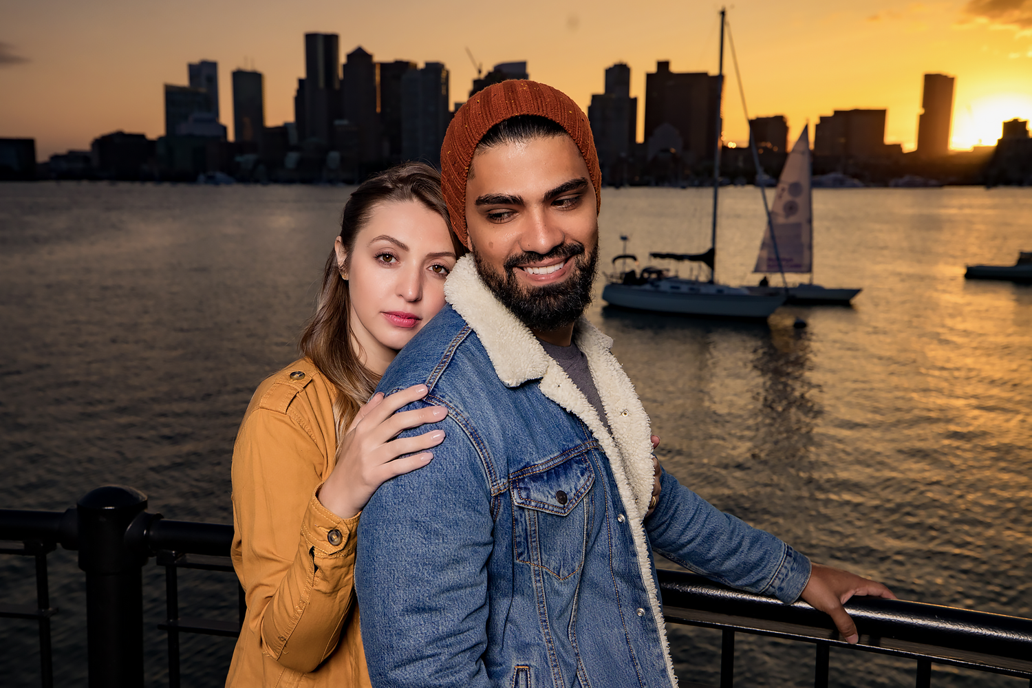 Couple poses together at sunset on a waterfront with sailboats and city skyline in the background.