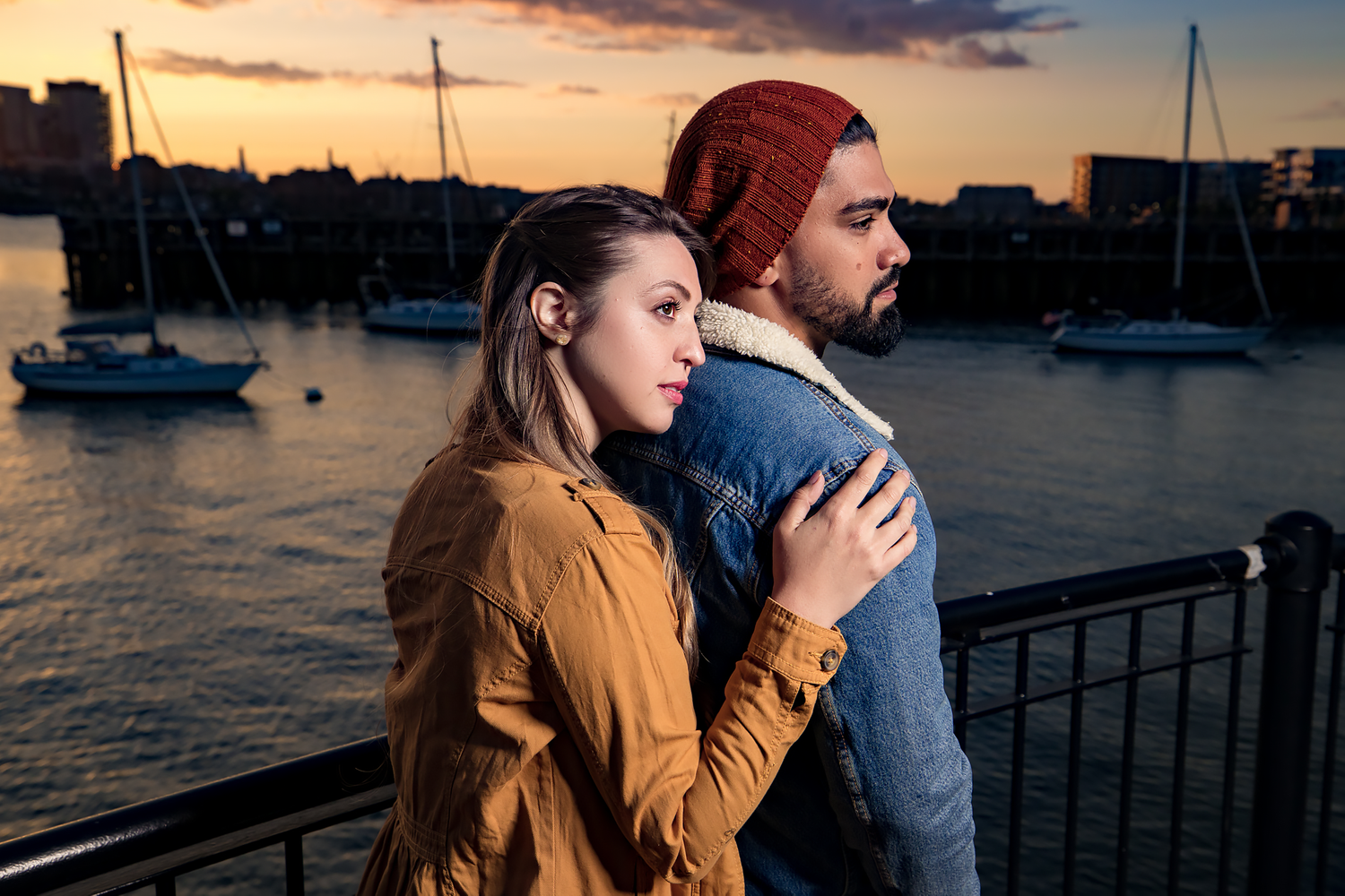Couple embraces on waterfront during sunset with boats in harbor behind them.