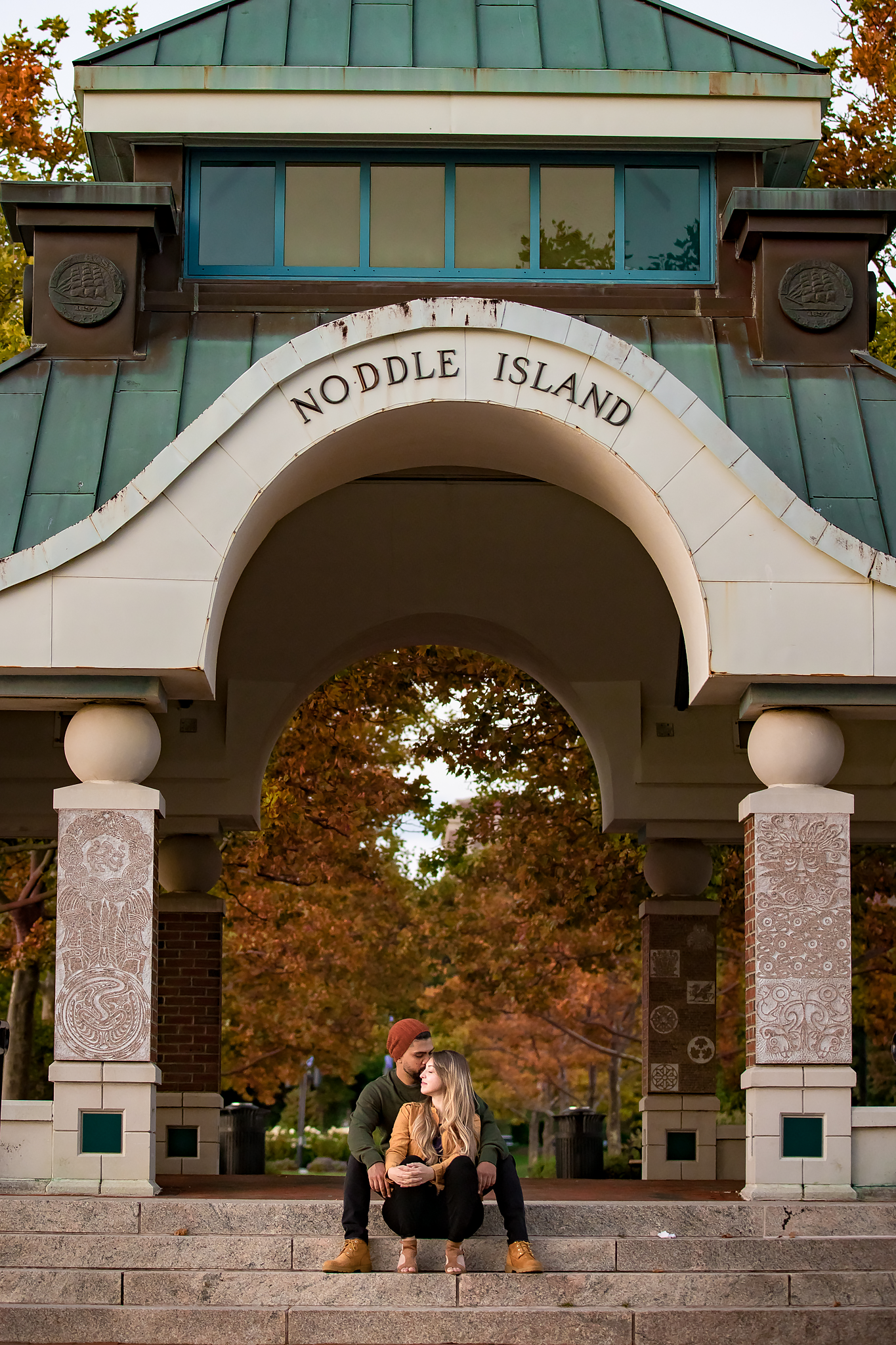 Couple sits beneath historic Noddle Island archway surrounded by autumn foliage.