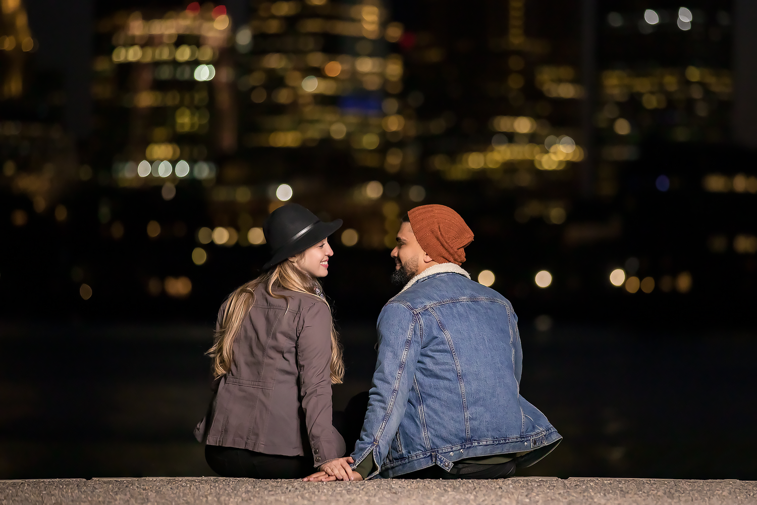 Couple sits together at night overlooking city lights reflected on dark water.
