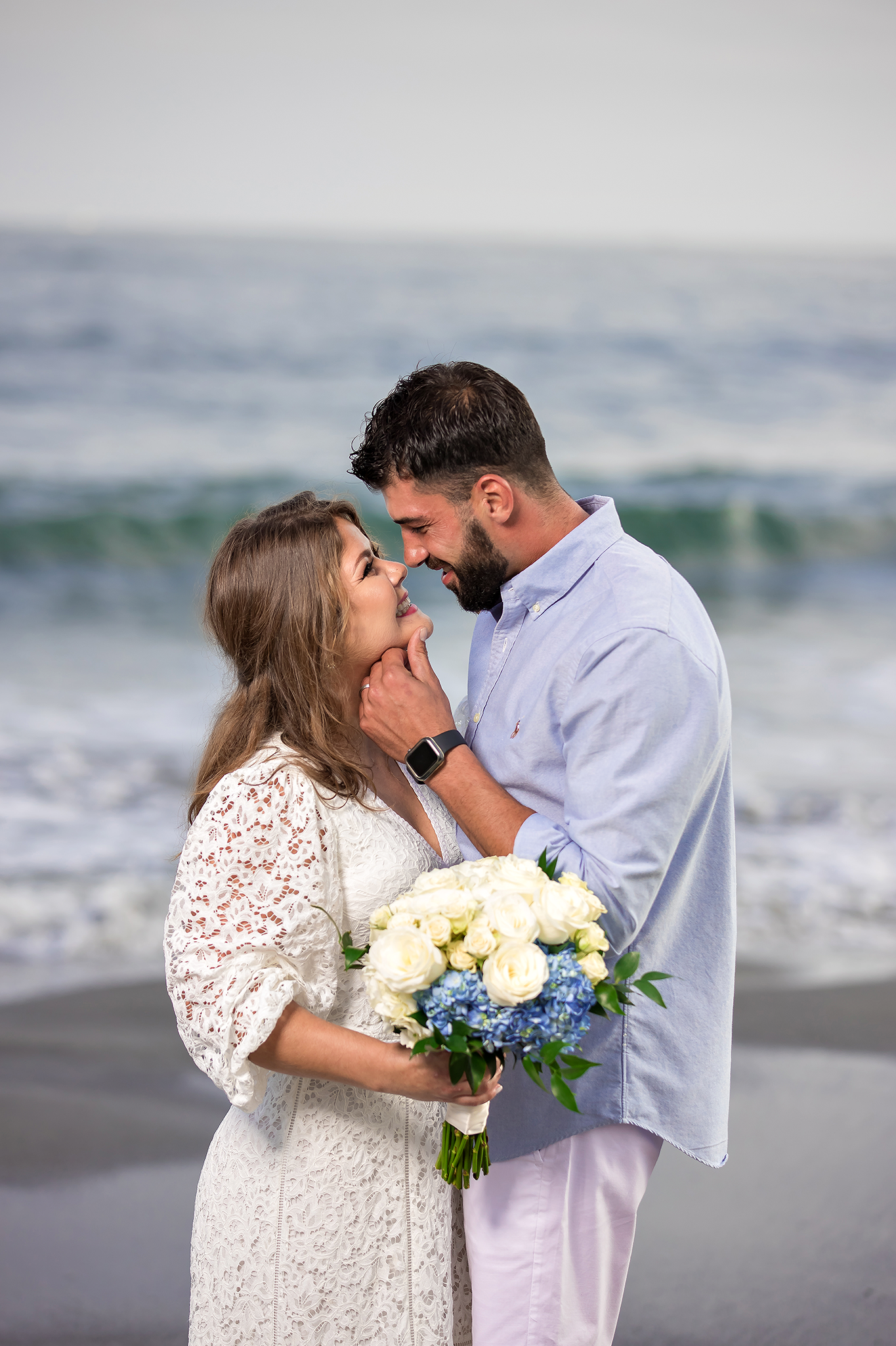 A couple shares an intimate moment on a beach with white roses and ocean waves behind them.