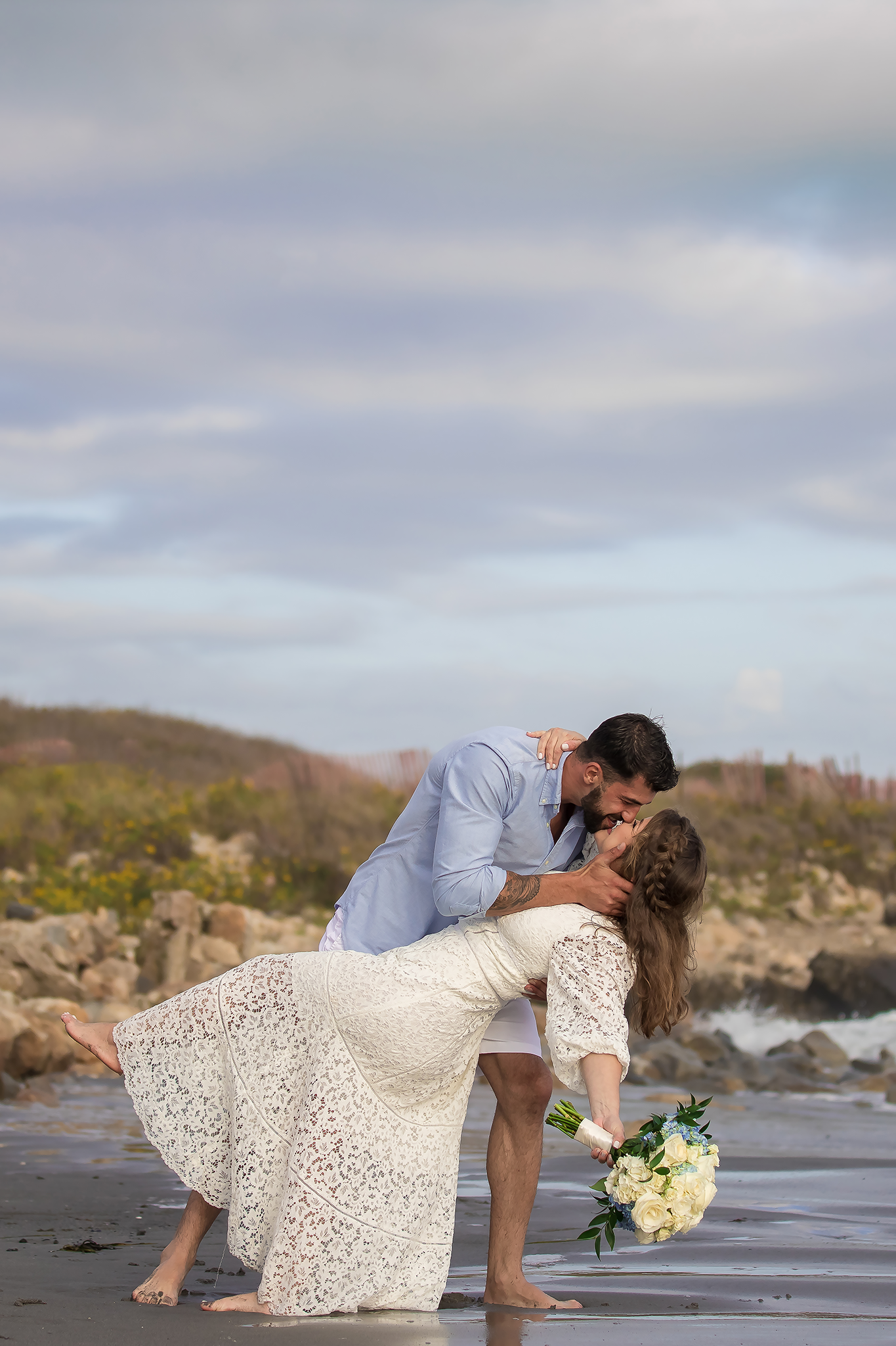 A romantic beach photo session showing a dip pose with a white lace dress against a rocky coastal backdrop.