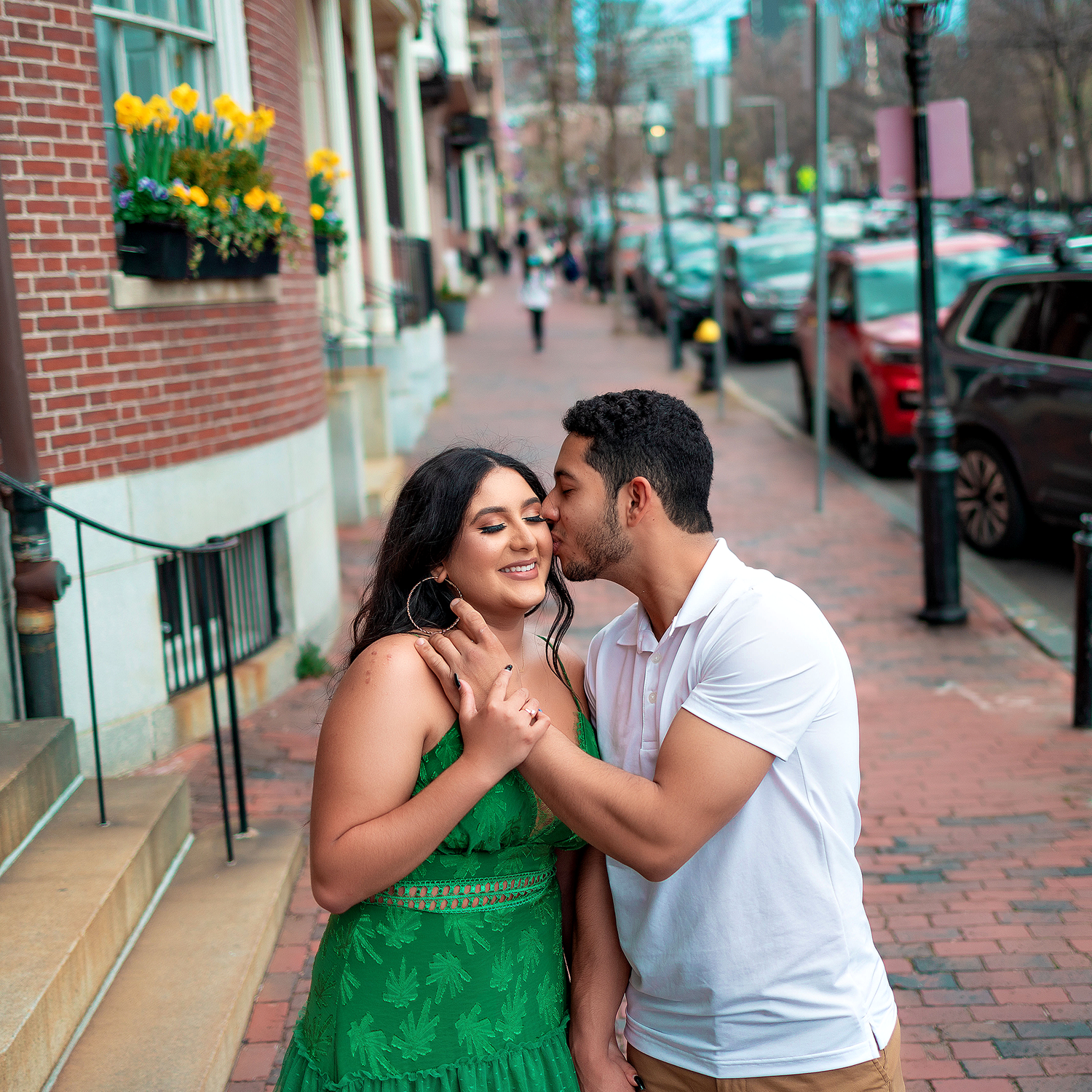 Couple sharing intimate moment on brick-lined street with yellow flowers in window box.