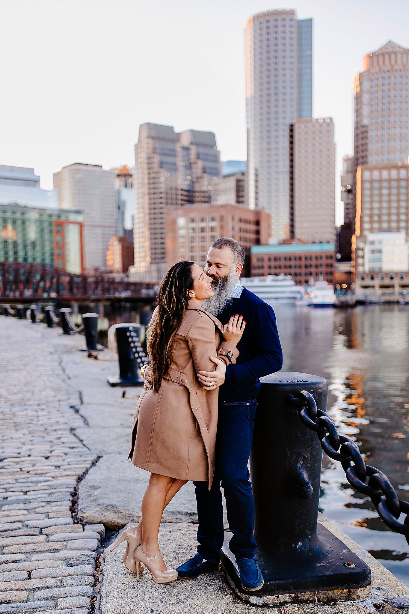 Couple embracing by waterfront with Boston harbor and city skyline at sunset.