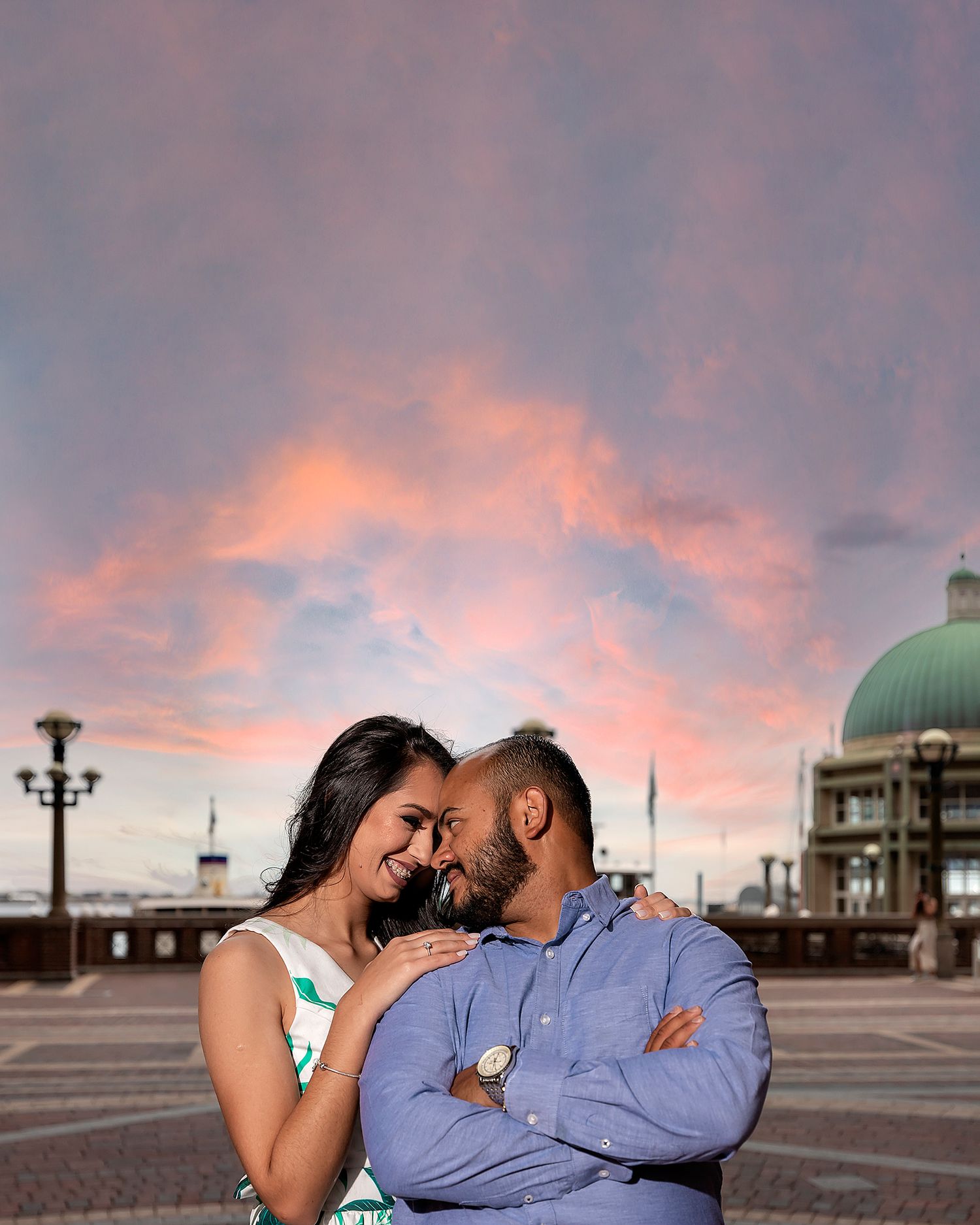 Romantic couple sharing an intimate moment against a pink sunset sky with a domed building in the background.