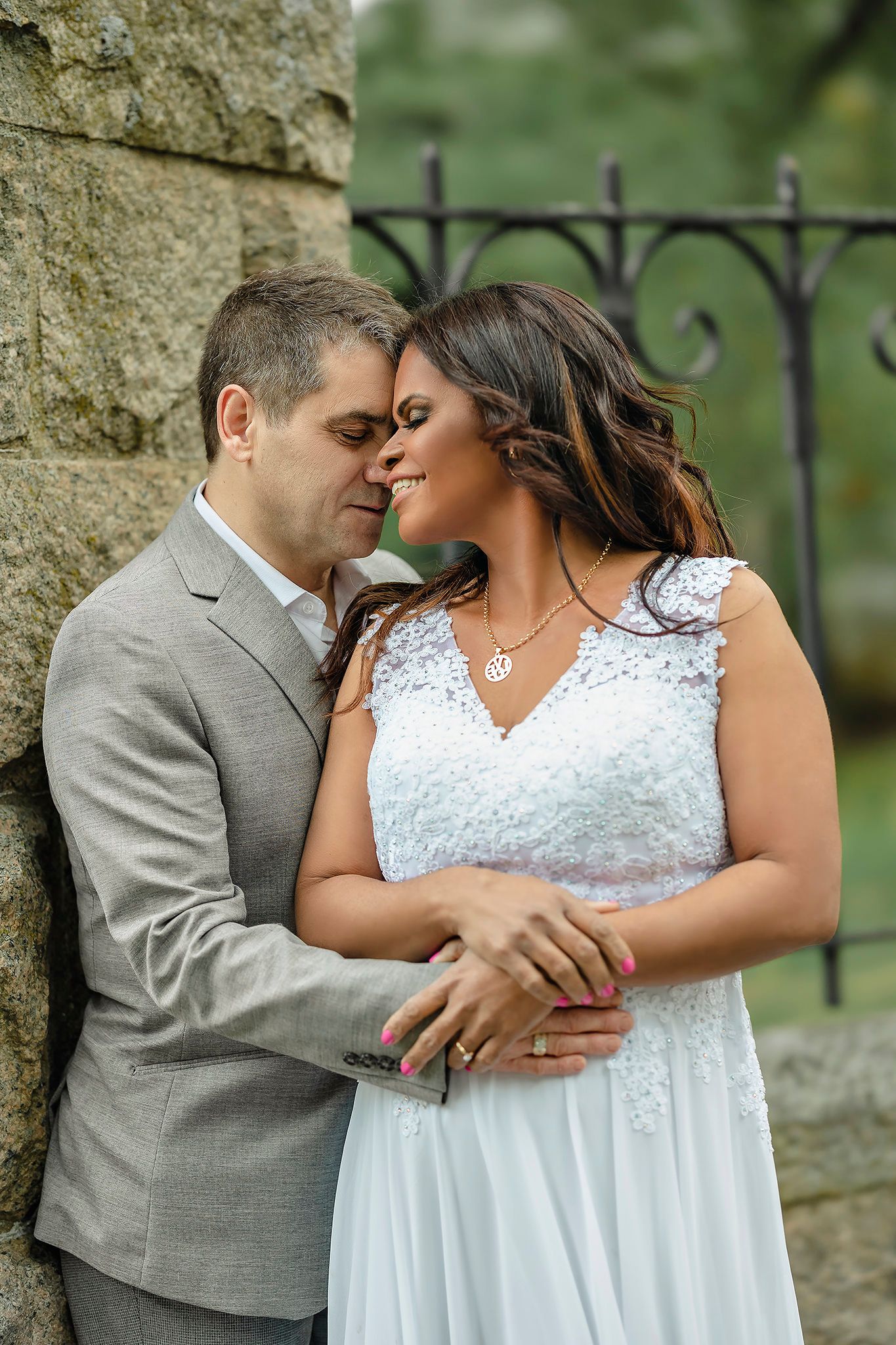 Romantic couple embraces against stone wall while wearing white dress and grey suit.