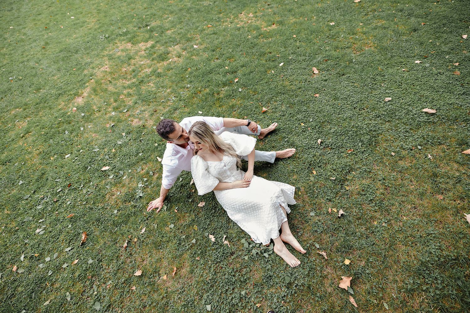 Couple in white outfits lying playfully on grass during photoshoot.