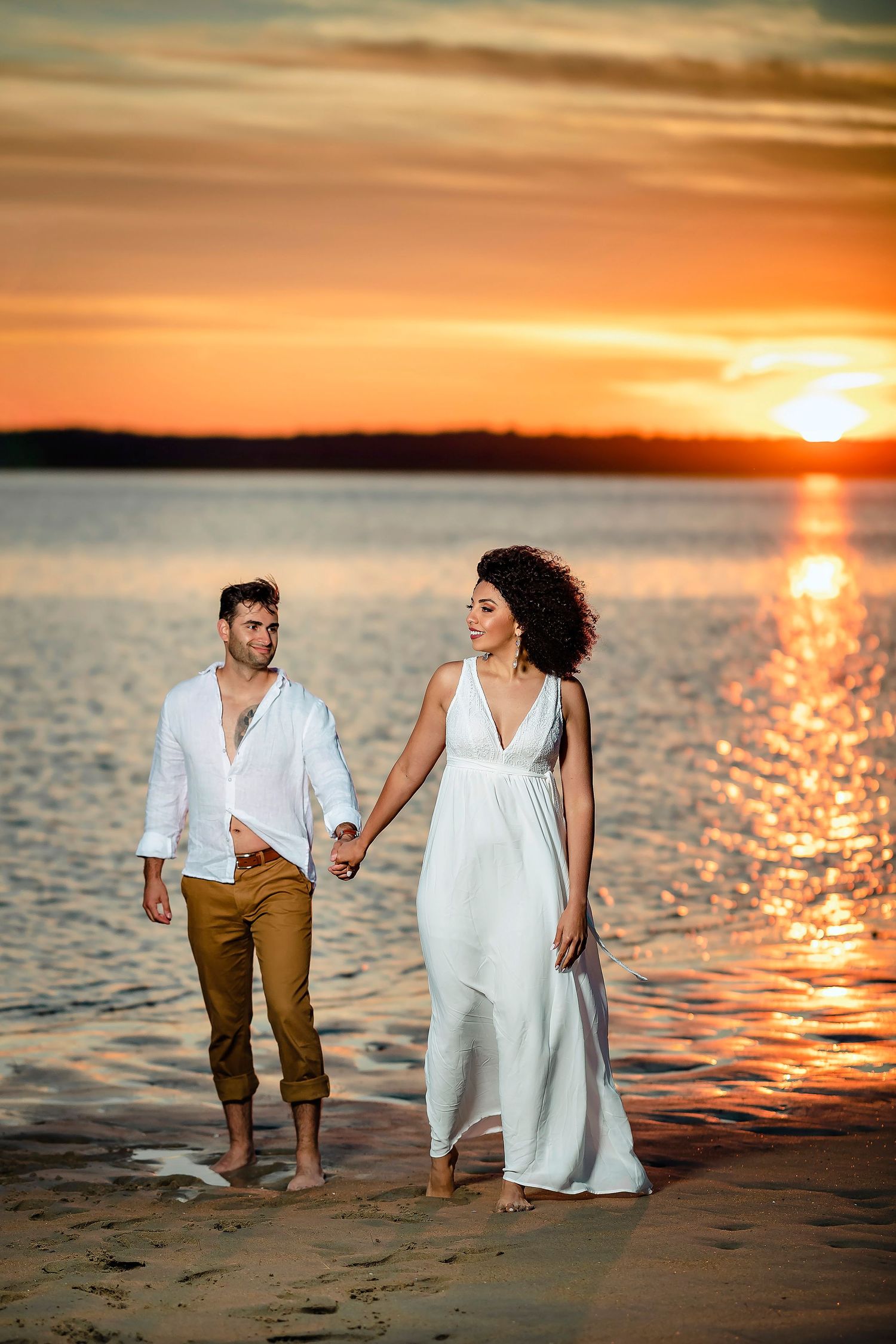 Couple walking along sandy beach at golden sunset with ocean waves sparkling.