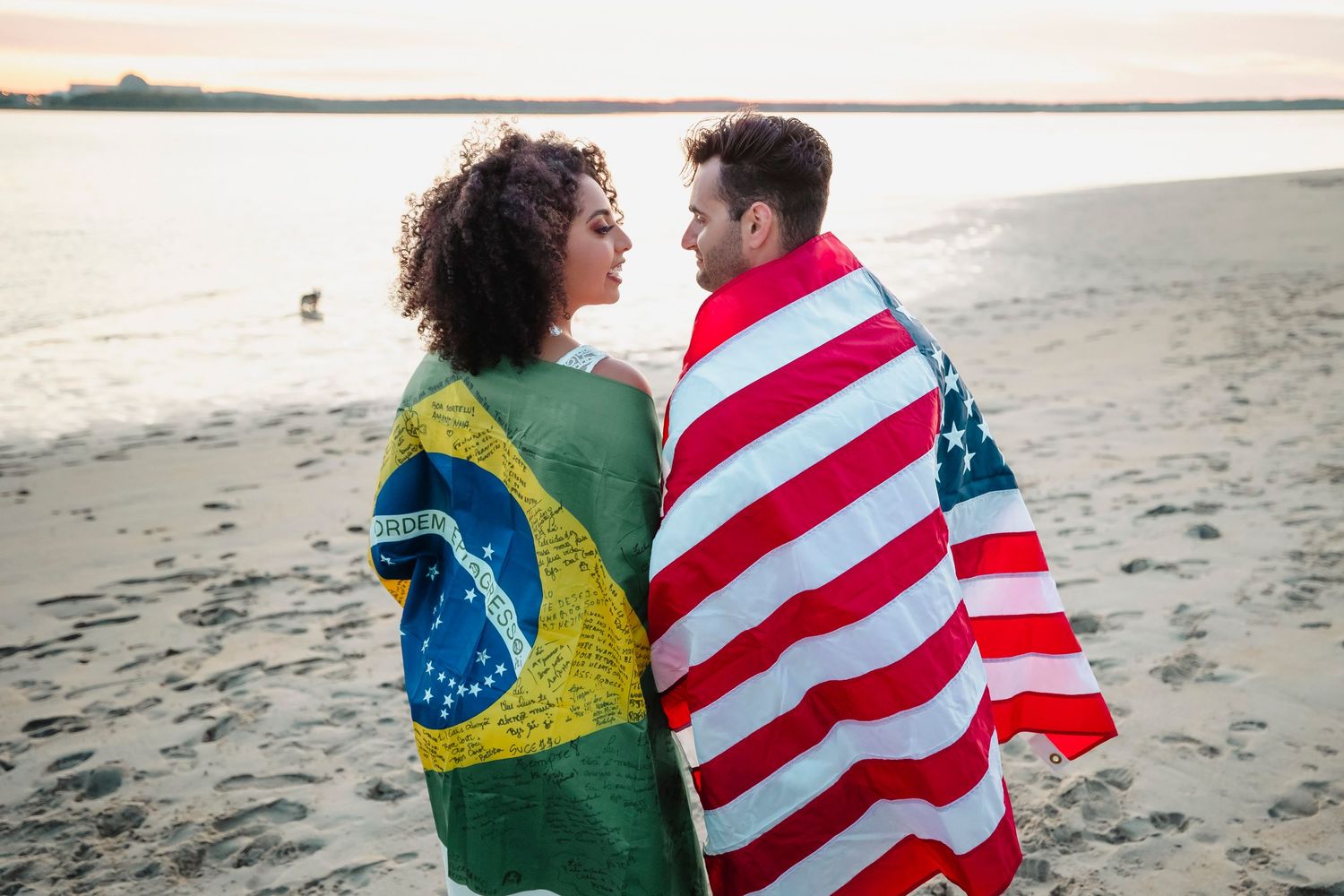Couple wrapped in American and Brazilian flags embrace on beach at sunset.
