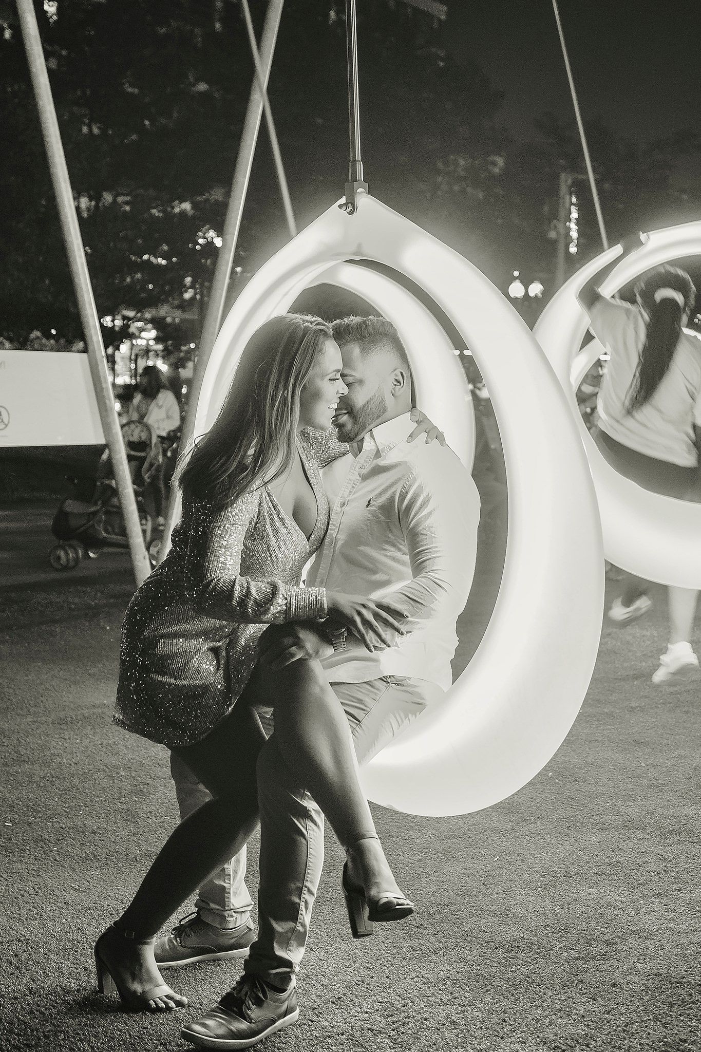 Black and white photo of playful couples on outdoor swings against a dark background.