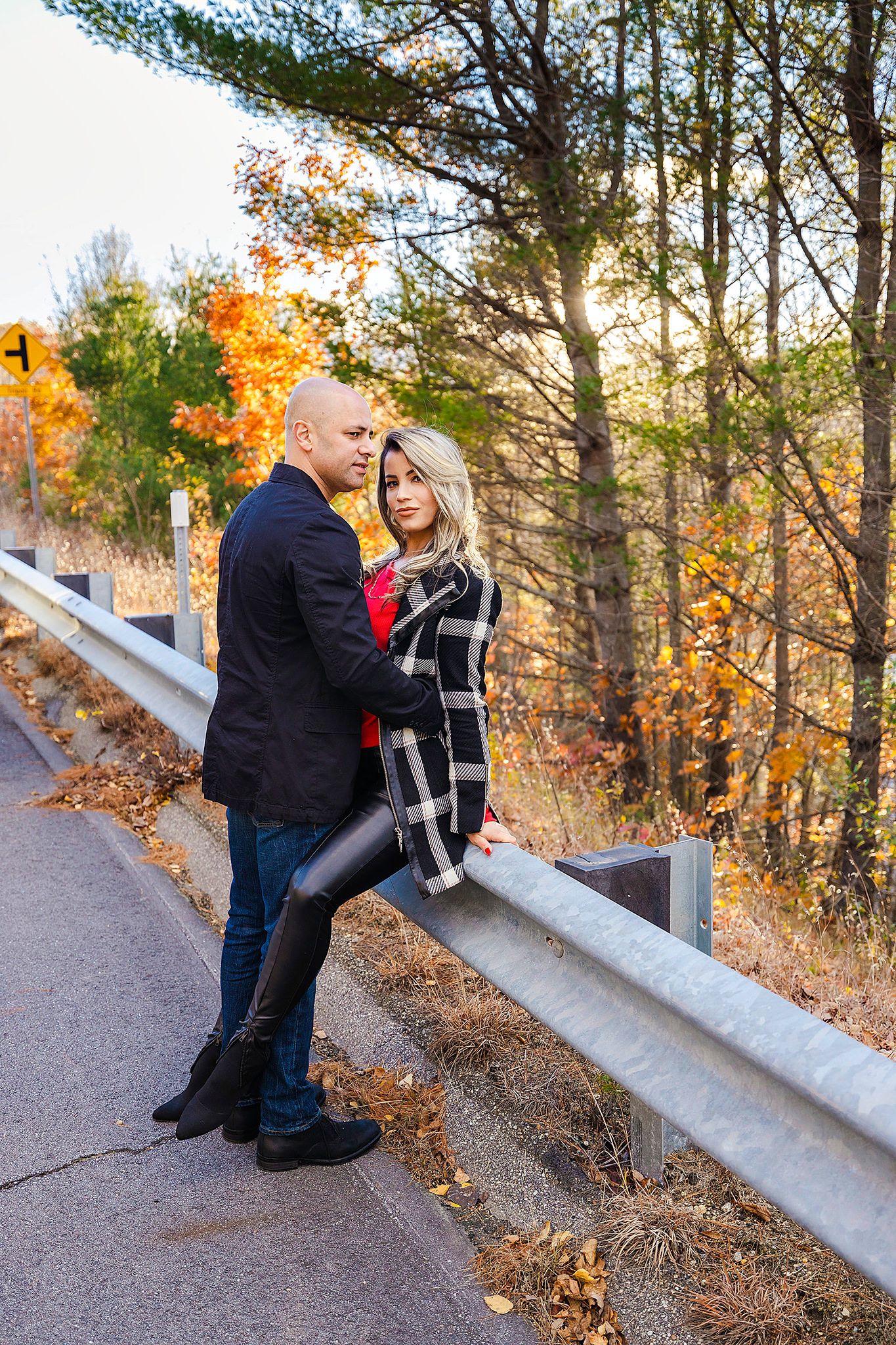 A couple poses romantically against a guardrail with fall foliage and sunlight in the background.