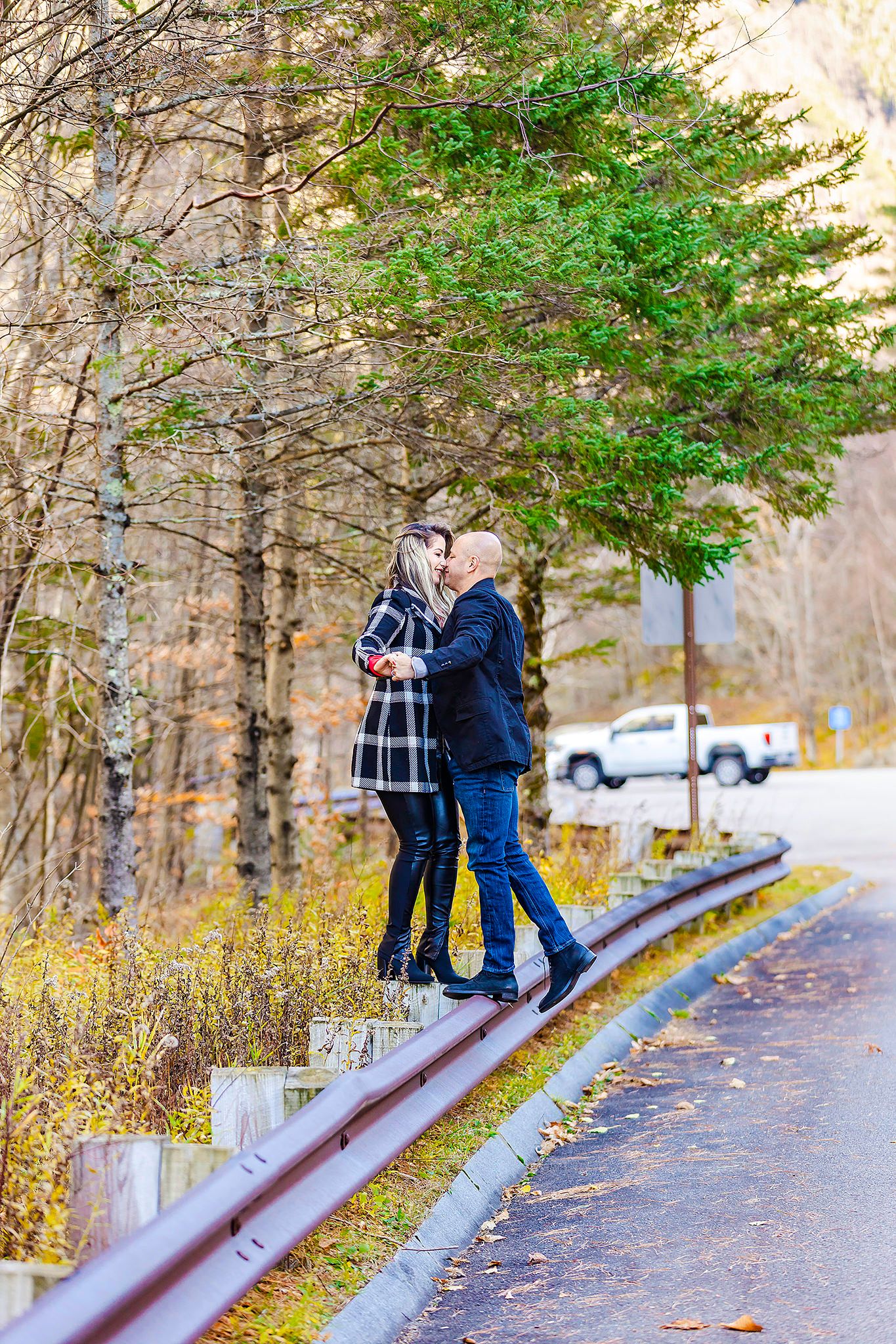 A couple stands together on a guardrail along a scenic mountain road in autumn.