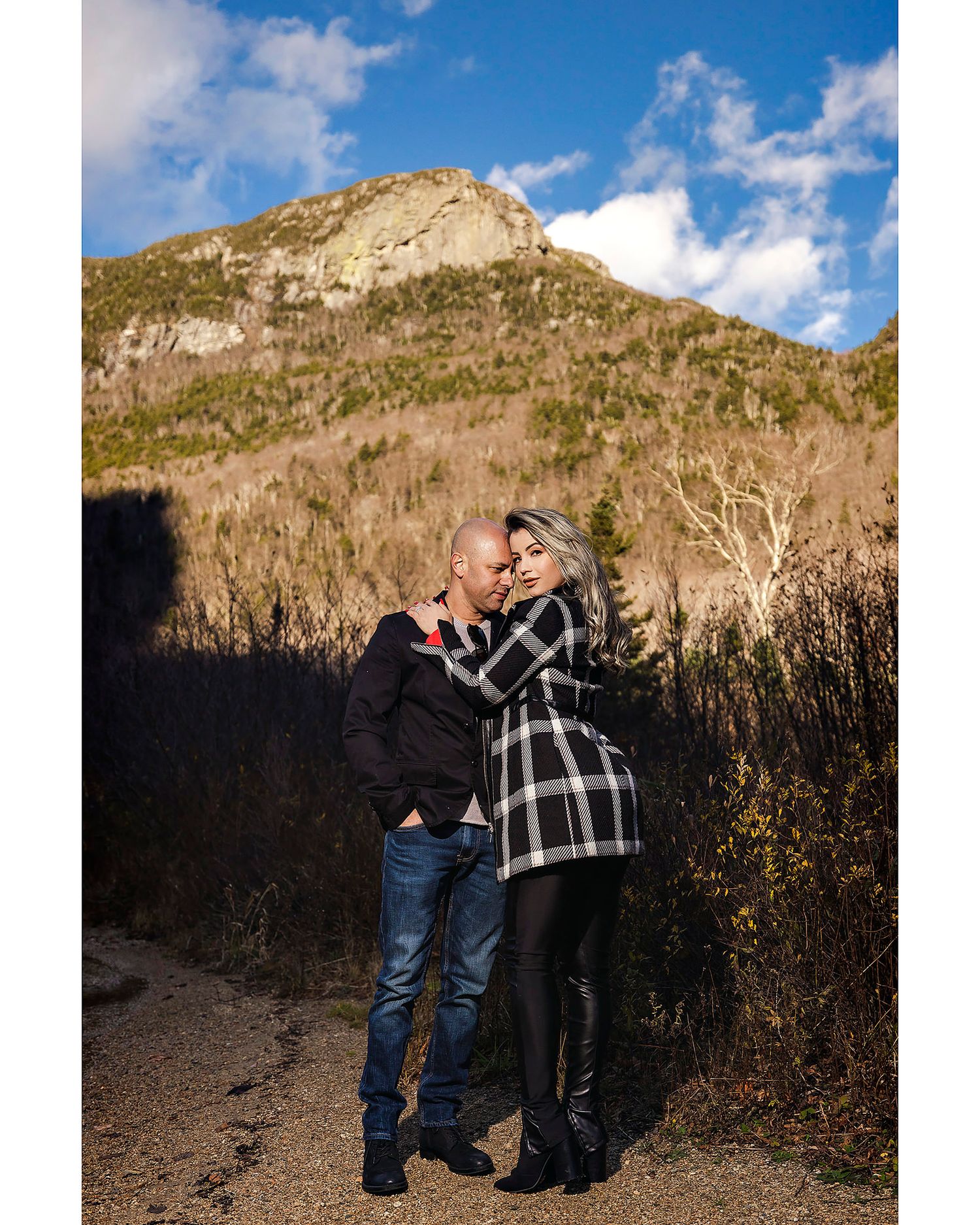 A couple embraces on a mountain trail with dramatic rocky peaks and blue sky in the background.