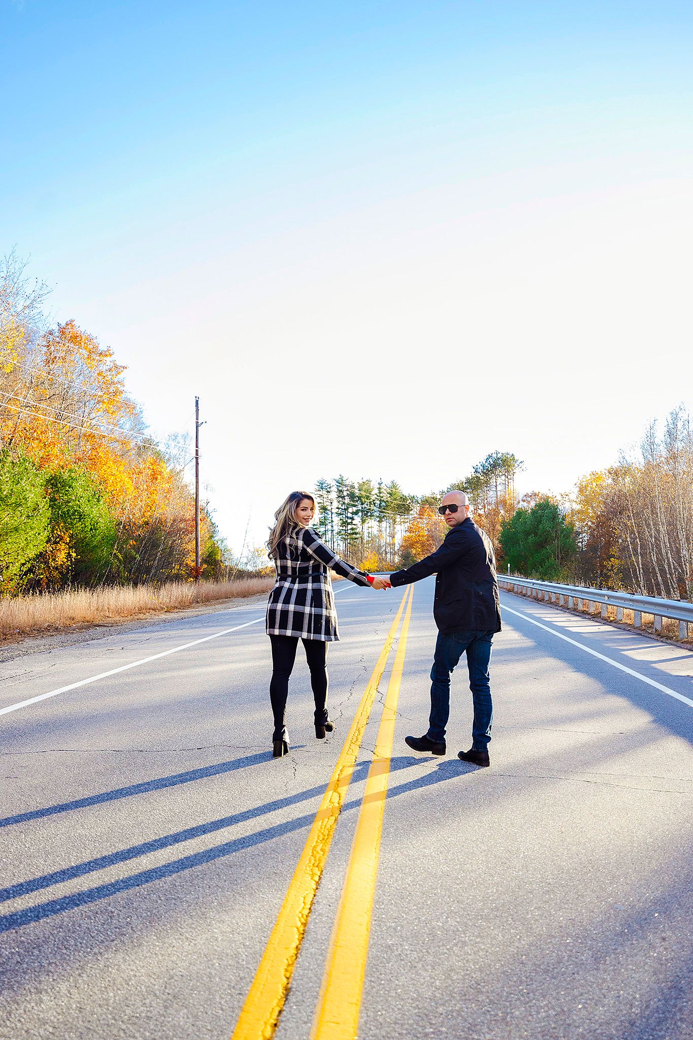 A couple holds hands while walking down the middle of an empty mountain road surrounded by autumn trees.