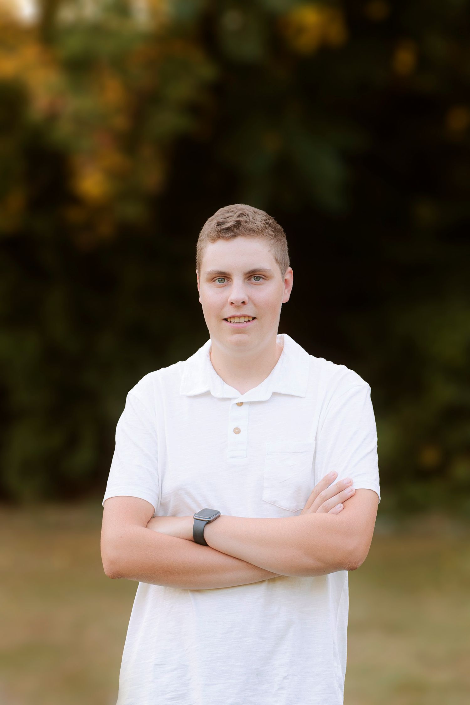 A casual outdoor portrait showing a person in a white polo shirt with crossed arms and a calm smile.