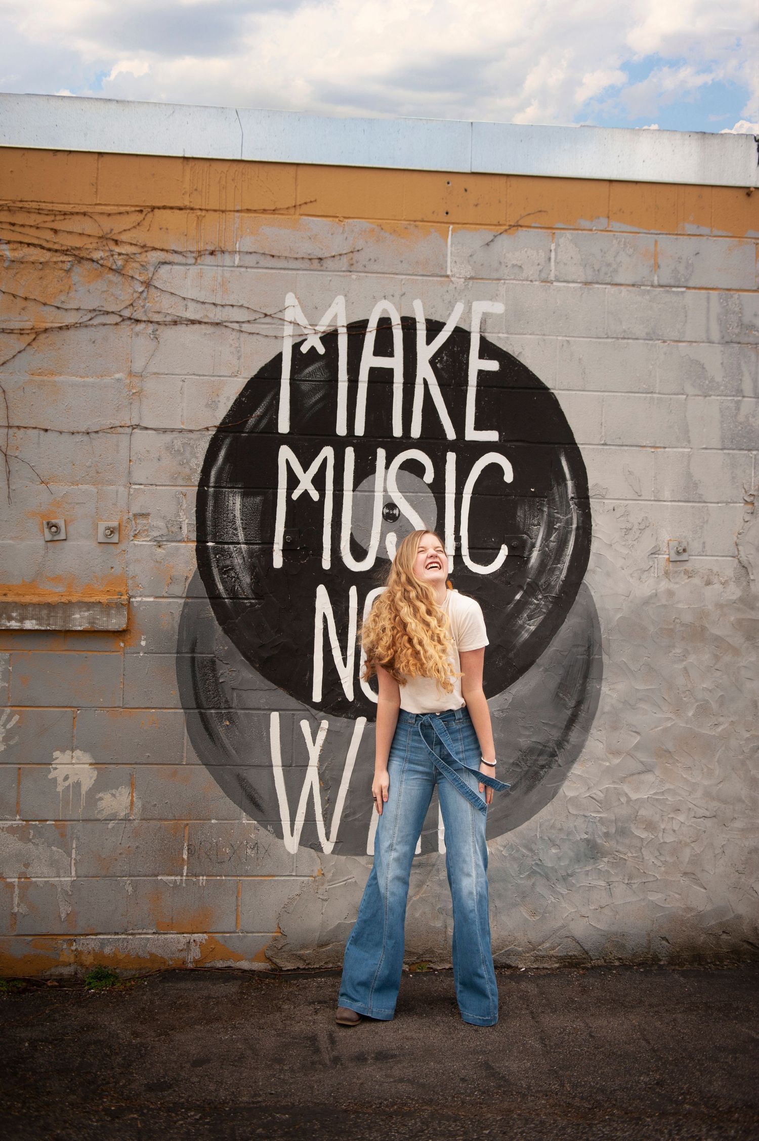 Person posing against wall with 'Make Music Now' graffiti art in black circular design.