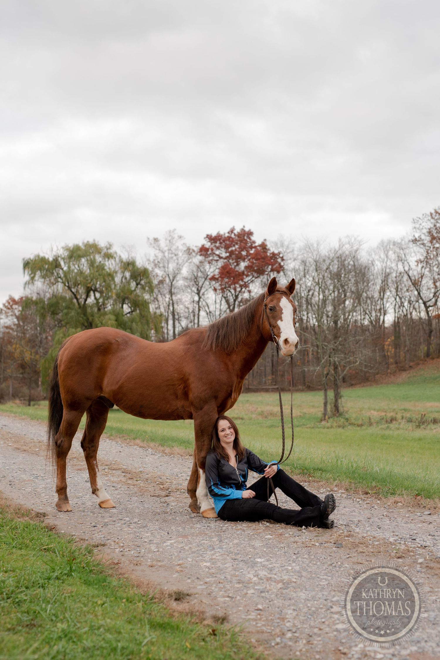 A chestnut horse stands attentively while someone sits on the ground of a country path.
