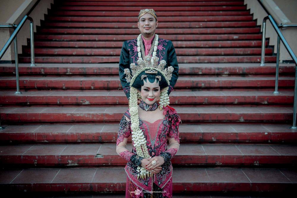 Traditional Indonesian wedding couple posing on red carpeted stairs in ceremonial attire.
