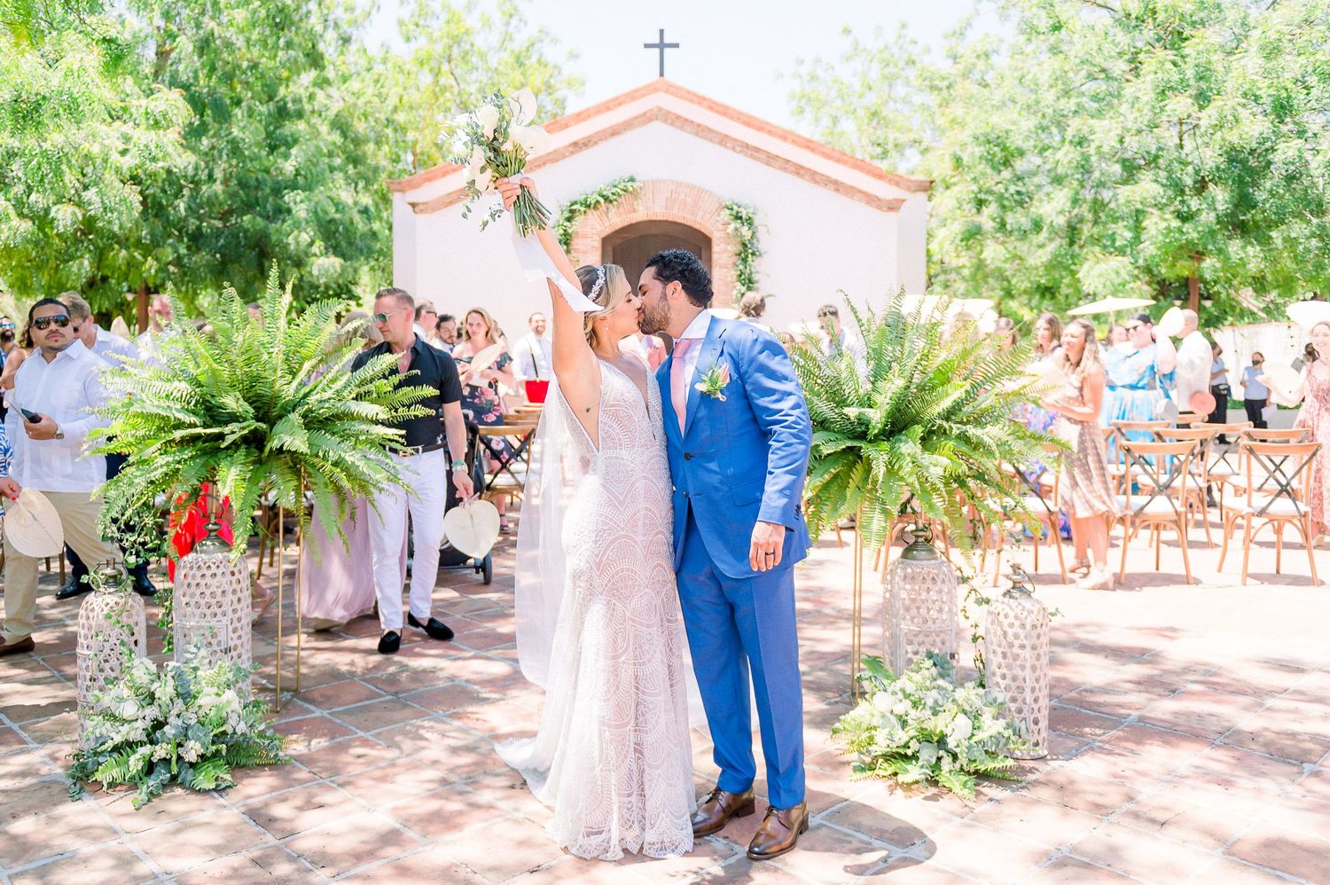 Newlyweds share their first kiss outside a charming chapel with tropical greenery and guests watching.