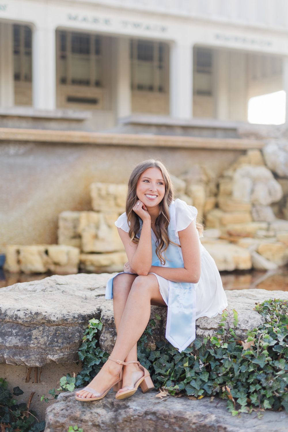 Woman in white summer dress sits on stone ledge near classic columned building.