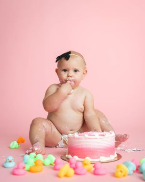 Baby girl smashes pink ombre birthday cake surrounded by colorful rubber ducks on pink background.