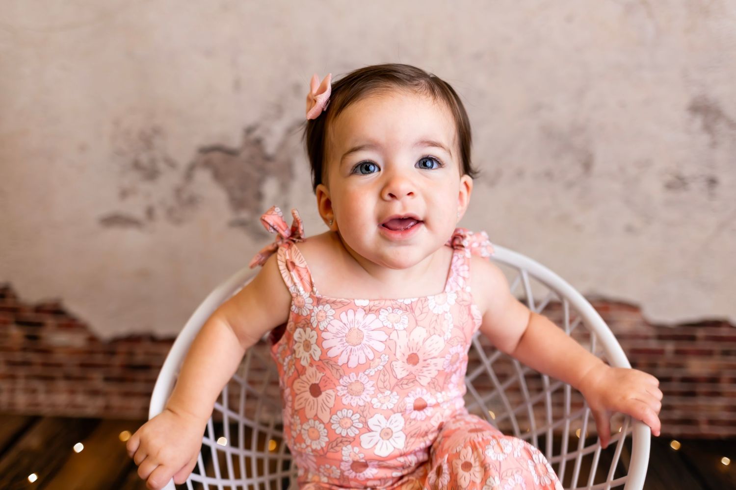 Joyful baby in pink outfit laughing while sitting in white chair against rustic backdrop.