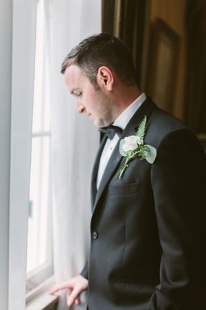 A formal portrait of a groom in a black tuxedo looking pensively out a window.