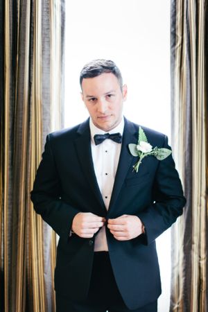 A formal portrait of a groom adjusting his bowtie in a window-lit room.