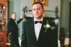 A classic groom portrait in a black tuxedo with white boutonniere against an interior backdrop.