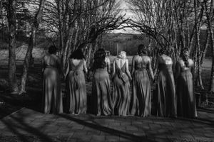 Black and white photo of bridesmaids in flowing dresses standing in a row among bare trees.