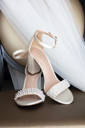 Elegant pearl-embellished white wedding sandals with block heels next to flowing tulle veil.
