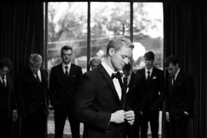 Groomsmen gather around the groom in formal attire by a window in black and white.