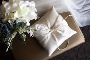 White silk ring bearer pillow with bow and floral decorations on dark surface.