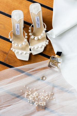 Pearl-embellished bridal shoes with veil, perfume bottle and hair accessories on wooden floor.