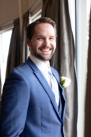 Portrait of person in blue suit with boutonniere by window with dark curtains.