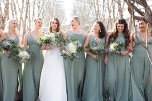 Bridal party in sage green dresses holding white bouquets while sharing a joyful moment outdoors.