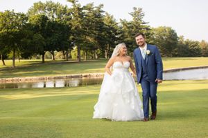 Newlyweds walking on golf course with water feature and trees in background.