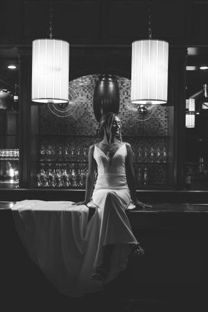 A dramatic black and white photo of a bride sitting at an elegant bar with vintage pendant lighting.