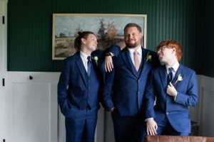 Three people in coordinated navy suits pose casually against a green wall with artwork.