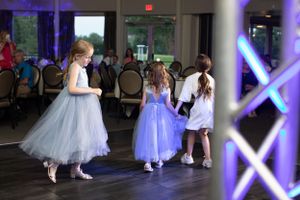 Young flower girls in tulle dresses dance on a reception floor illuminated by purple lights.