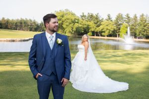 A navy blue suited groom stands on a manicured golf course with a pond and fountain view.