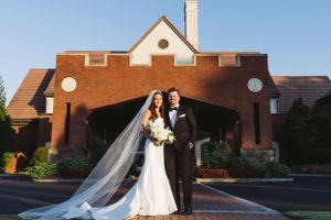 A wedding couple poses outside a red brick building with a long flowing veil on a sunny day.